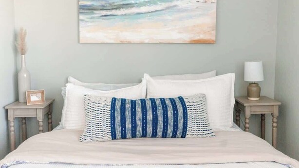 A bed with white bedding, a blue and white striped pillow, and a painting of a beach scene on the wall. - Newport's favorite Neighborhood 3bed (Newport)