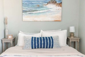 A bed with white bedding, a blue and white striped pillow, and a painting of a beach scene on the wall.