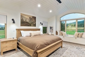 A bedroom with a large bed, wooden headboard, and a view of a green landscape through a large window.