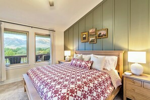 A bedroom with a large bed, two bedside lamps, a view of mountains through the windows, and a wall with framed pictures.