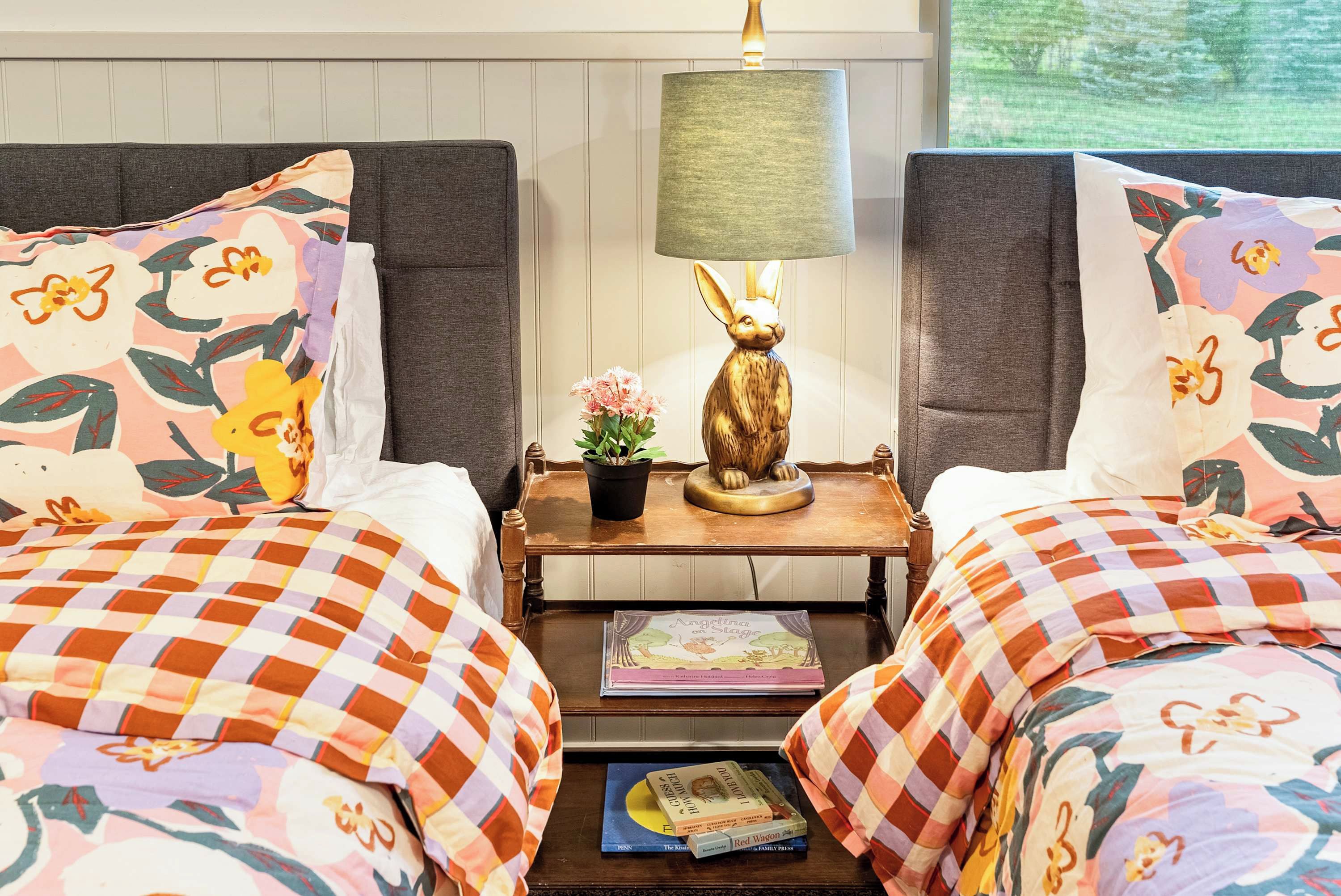A hotel room with two beds, a nightstand with a lamp and a bunny figurine, and a window with a view of trees.