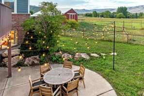 An outdoor patio with a round wooden table and chairs, surrounded by a stone wall and string lights.