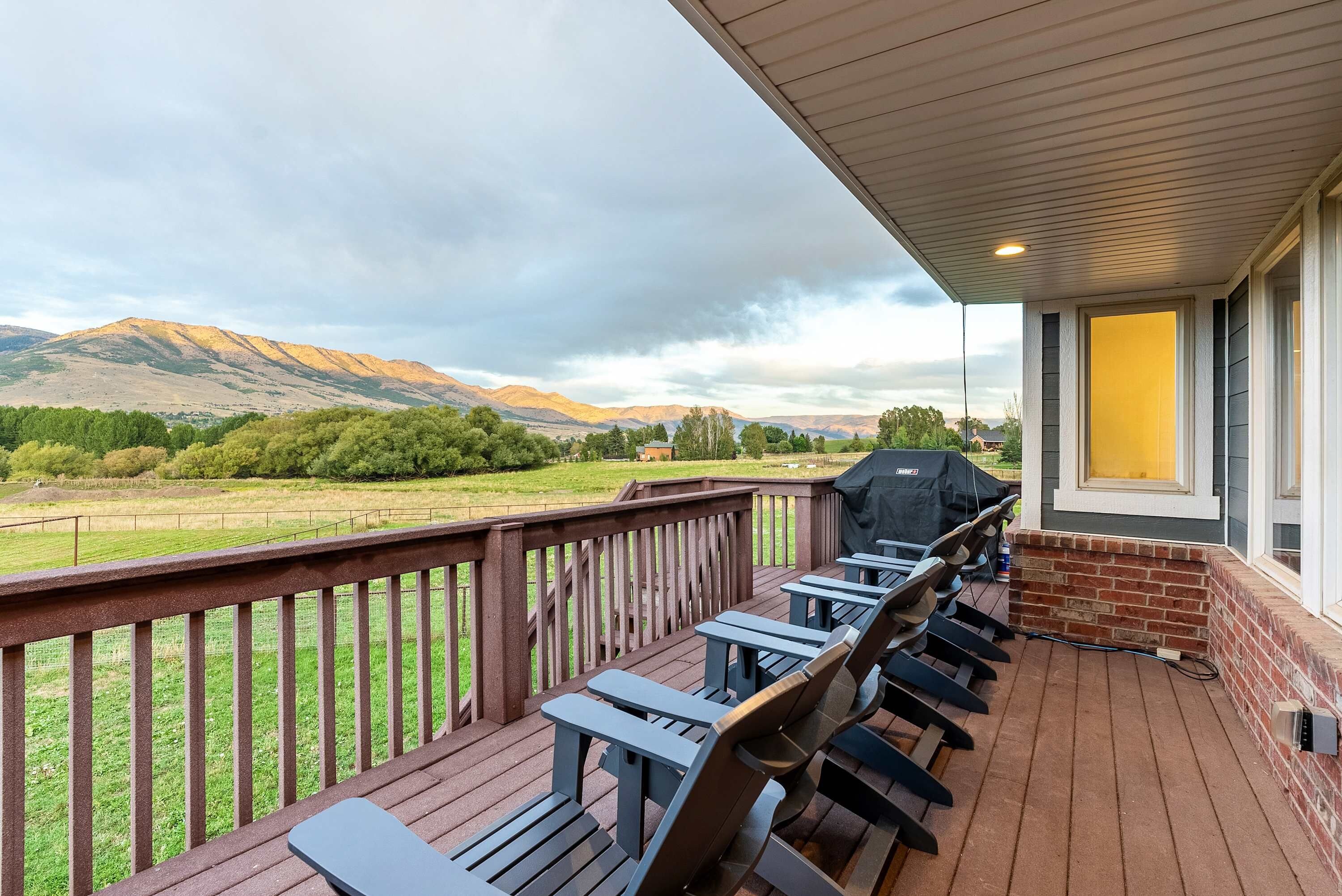 A covered outdoor deck with a barbecue grill, wooden chairs, and a view of a mountainous landscape.