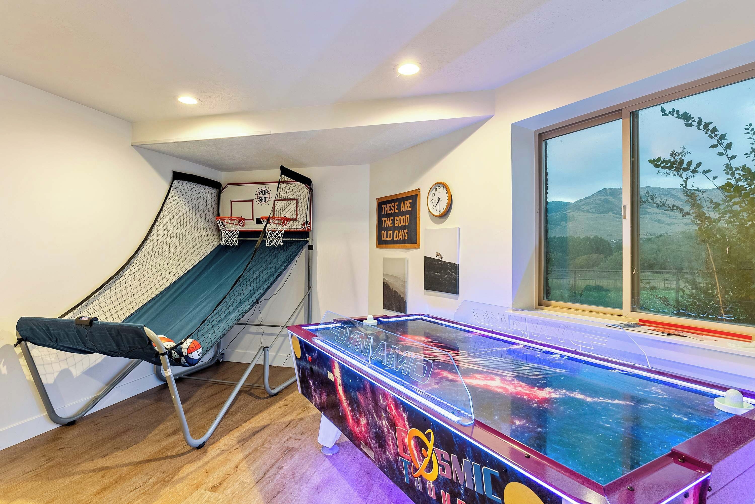 A room with a ping pong table, a basketball hoop, and a large window with a view of hills.