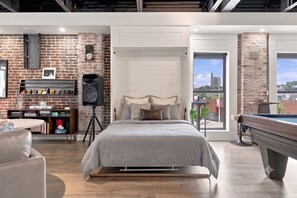 Une chambre moderne avec un mur en briques, un lit avec de nombreux oreillers, un haut-parleur, une table de billard et une vue sur la ville.