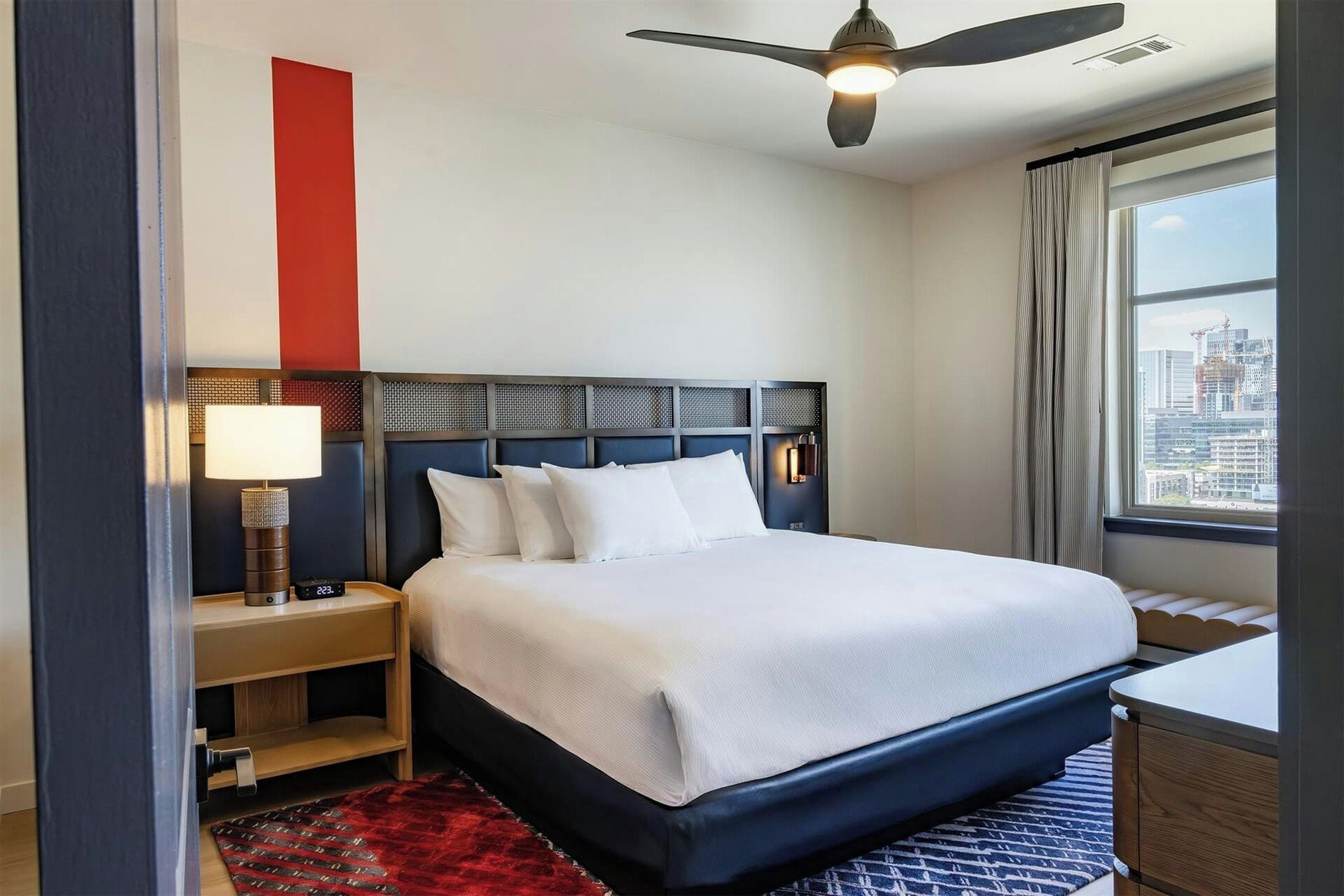 Una habitación de hotel con cama, mesita de noche, lámpara, ventilador de techo y ventana con vista a la ciudad.