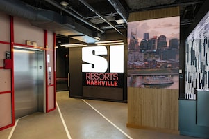 Um saguão de hotel com elevador, uma grande placa iluminada com a inscrição 'RESORTS NASHVILLE' e vista para a paisagem urbana na parede.