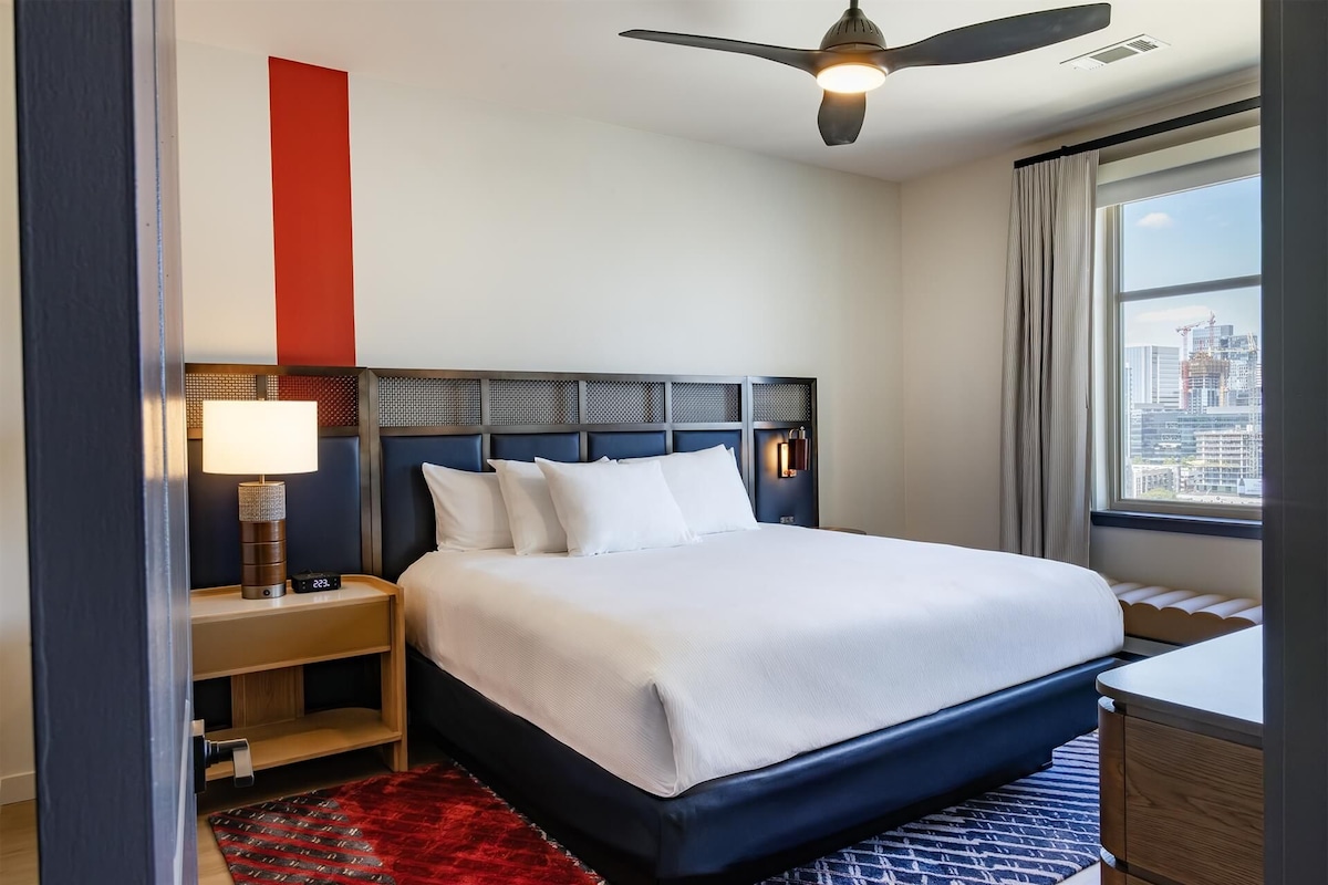 Ein Hotelzimmer mit Bett, Nachttisch, Lampe, Deckenventilator und Fenster mit Stadtblick.