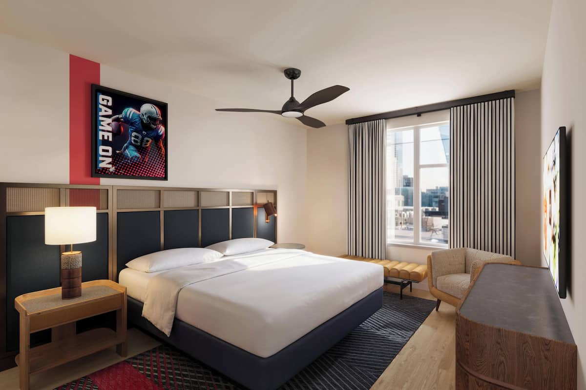 A hotel room with a bed, a desk, a chair, a ceiling fan, and a window with curtains.