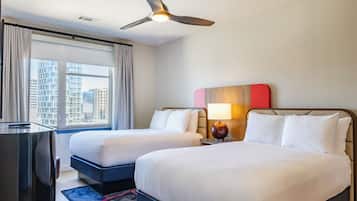 A hotel room with two beds, a desk, a ceiling fan, and a view of the cityscape.