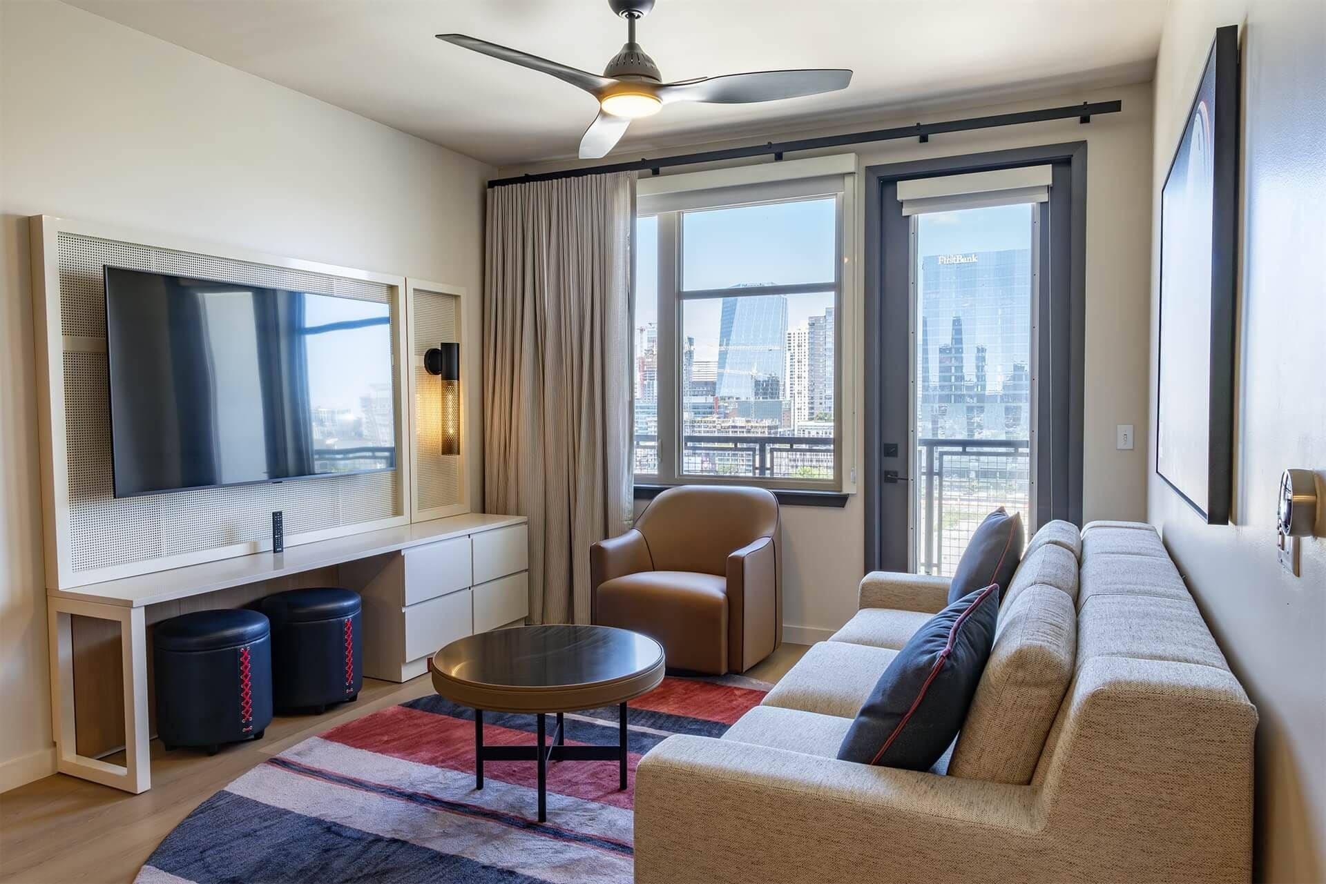 Una sala de estar moderna con un televisor de pantalla plana grande, un ventilador de techo, un sofá, una silla y vistas de la ciudad a través de amplias ventanas.