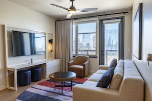 Una sala de estar moderna con un televisor de pantalla plana grande, un ventilador de techo, un sofá, una silla y vistas de la ciudad a través de amplias ventanas.
