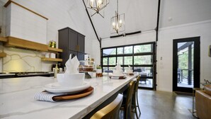 A modern kitchen with a long island, wooden chairs, and a view of the outdoors through large windows. - Couples & Kids Barndominium Style Retreat (Broken Bow)