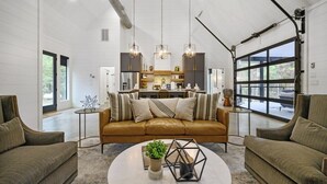 A spacious living room with a brown leather sofa, two armchairs, a round coffee table, and pendant lighting. A kitchen is visible in the background with a large window. - Couples & Kids Barndominium Style Retreat (Broken Bow)
