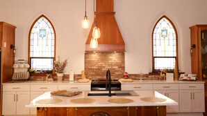Une cuisine dotée d’un îlot central, de deux grandes fenêtres ornées de vitraux, d’une cheminée en briques et de meubles de rangement en bois.