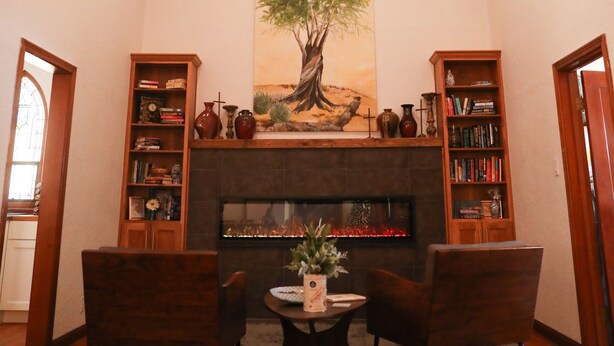 Une pièce chaleureuse avec une cheminée, des étagères en bois remplies de livres, deux fauteuils et un grand tableau représentant un arbre accroché au mur.