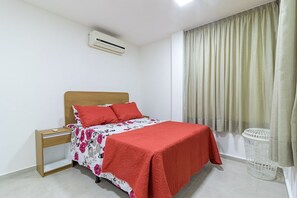 Ein Schlafzimmer mit einem Bett, einem Nachttisch, einer Klimaanlage, einem Fenster mit Vorhängen und einem Wäschekorb.