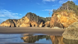 A coastal landscape with rocky cliffs, a sandy beach, and a calm water body reflecting the sky. - Oceanside Escape • An Oceanfront Gathering Place (Arch Cape)