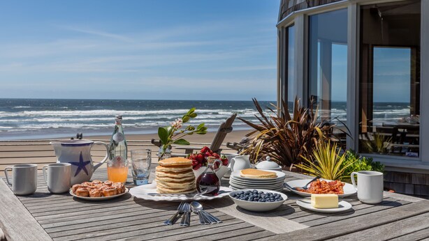 A breakfast table with a view of the ocean, featuring pancakes, fruit, and a bottle of juice. - Oceanside Escape • An Oceanfront Gathering Place (Arch Cape)
