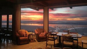 A cozy interior with leather chairs and a dining table, large windows offering a view of the beach and sunset. - Oceanside Escape • An Oceanfront Gathering Place (Arch Cape)
