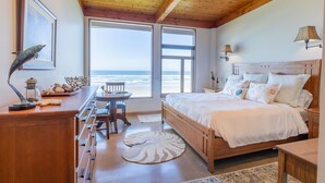A bedroom with a wooden ceiling, a large bed with white bedding, a desk with a lamp, and a view of the ocean through large windows. - Oceanside Escape • An Oceanfront Gathering Place (Arch Cape)