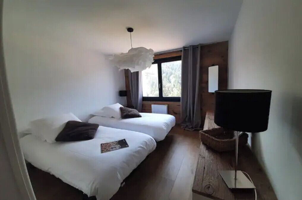 A bedroom with two beds, a window with curtains, a hanging light fixture, and a bedside table with a lamp.