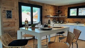 Une cuisine moderne avec des murs en bois, une table à manger blanche, des chaises en osier et une vue sur l’extérieur.