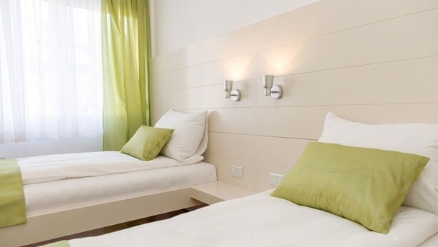 Ein Hotelzimmer mit zwei Betten, jedes mit weißer Bettwäsche und grünen Kissen. An der Wand über den Betten befinden sich Wandleuchten.