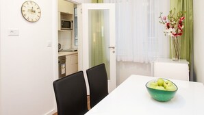 Ein Essbereich mit einem weißen Tisch, schwarzen Stühlen, einer grünen Schüssel mit Äpfeln, einer Wanduhr und einer Küche im Hintergrund.