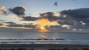 A beach at sunset with the sun setting over the ocean, clouds in the sky, and a person walking on the beach. - Direct Oceanfront  South Cocoa Beach. Watch launches from Balcony. (Cocoa Beach)