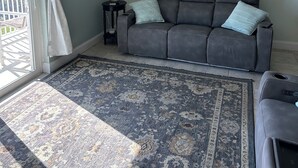 A living room with a grey sofa, patterned rug, a painting of beach chairs, and a ship's wheel on the wall. - Direct Oceanfront  South Cocoa Beach. Watch launches from Balcony. (Cocoa Beach)