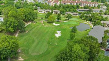 An aerial view of a golf course with a clubhouse, surrounded by residential buildings and lush greenery.