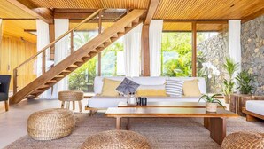 A modern living room with a wooden staircase, a sofa set, wicker ottomans, and a stone wall.