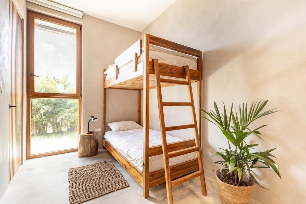 Un lit superposé avec une échelle en bois, un tapis tissé, une plante en pot et une vue sur la verdure par une grande fenêtre.