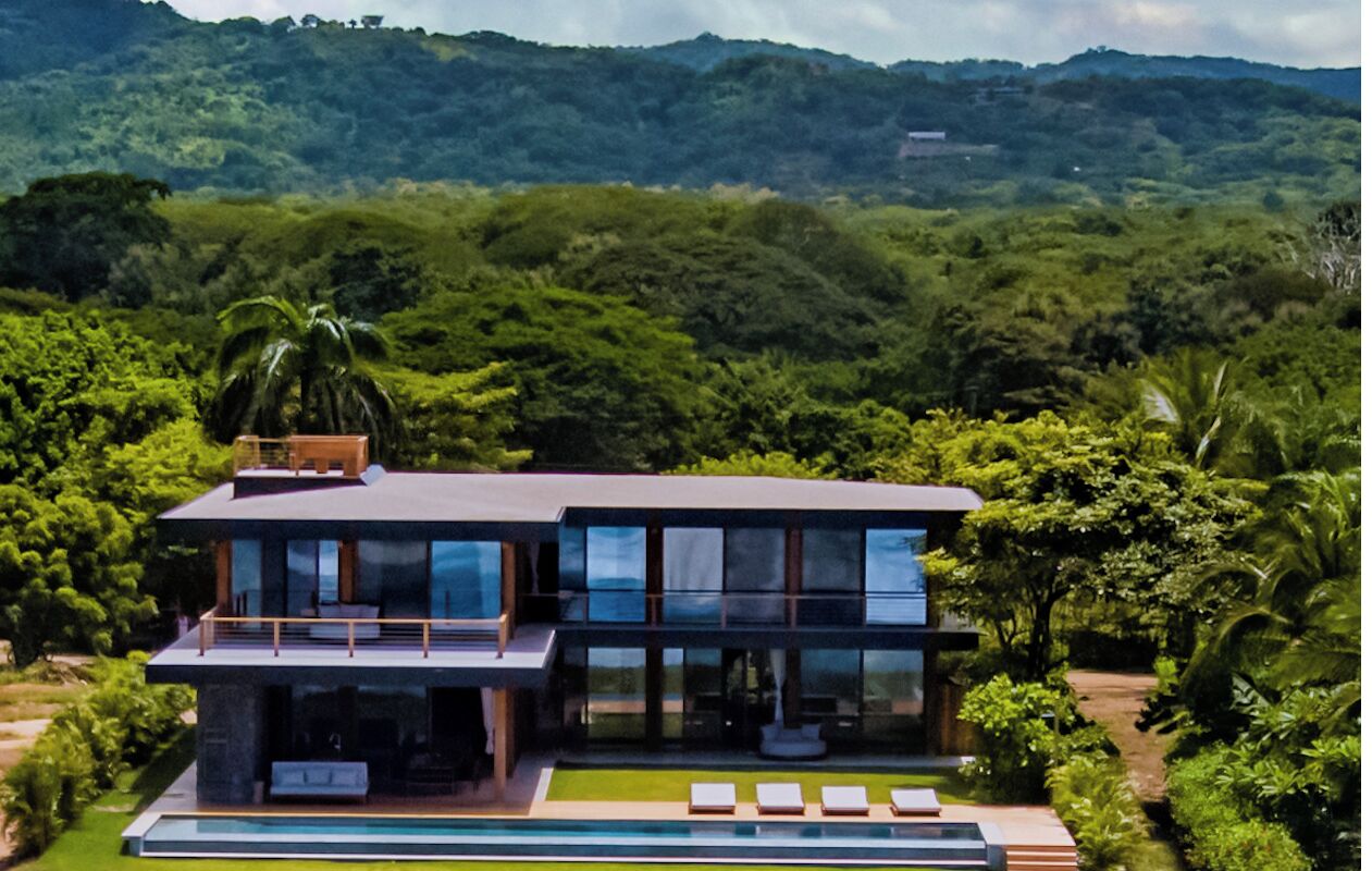 Une maison moderne avec une piscine, entourée d’une végétation luxuriante et de collines.