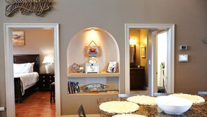 A hotel room with a bed, a desk with a lamp, a dining table with placemats, a wall-mounted shelf with decorative items, and a hallway leading to a bathroom. - East Camptons-Lakehouse And Pool (New Orleans)