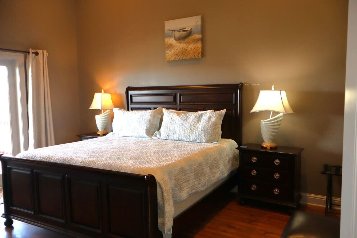 A neatly made bed with a dark wooden headboard, two bedside lamps, and a painting above the headboard.