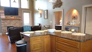 A kitchen with granite countertops, wooden cabinets, and a central island with a bowl. There is a trash can and a seating area with a leather couch. - East Camptons-Lakehouse And Pool (New Orleans)