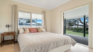 A bedroom with a large bed, a bedside table with a lamp, and a view of a residential area through the window. - Birdsong - Central location (Wooli)