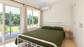 Un dormitorio con cama, ventana con cortinas, equipo de aire acondicionado y vista a zonas verdes.