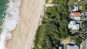 An aerial view of a coastal property with a sandy beach, dense greenery, and several modern houses with flat roofs. - Birdsong - Central location (Wooli)
