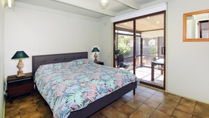 Une chambre à coucher comprenant un lit, deux lampes de chevet, une table de nuit en bois, une fenêtre donnant sur l’extérieur et une porte-fenêtre coulissante qui mène à un patio.