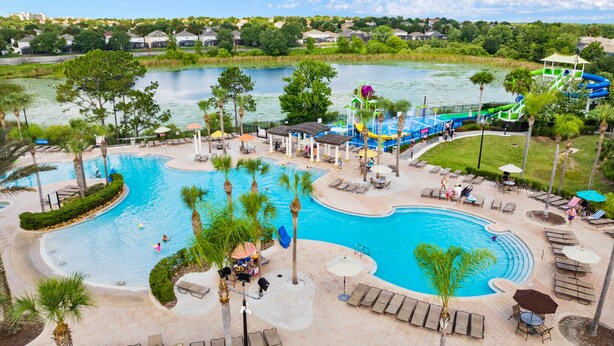 A large outdoor swimming pool with a water slide and lounge areas, surrounded by palm trees and a view of a lake. - 4BR Disney | Windsor Hills Resort | Pool Villa (Kissimmee)