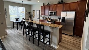 A modern kitchen with a central island, wooden cabinets, stainless steel appliances, and a dining area with chairs. - 4BR Disney | Windsor Hills Resort | Pool Villa (Kissimmee)
