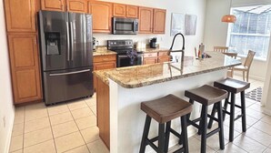 A modern kitchen with wooden cabinets, stainless steel appliances, a granite countertop, and a bar area with stools. - Disney | Pool&Spa | Windsor Hills | Privacy | Amazing Views (Kissimmee)