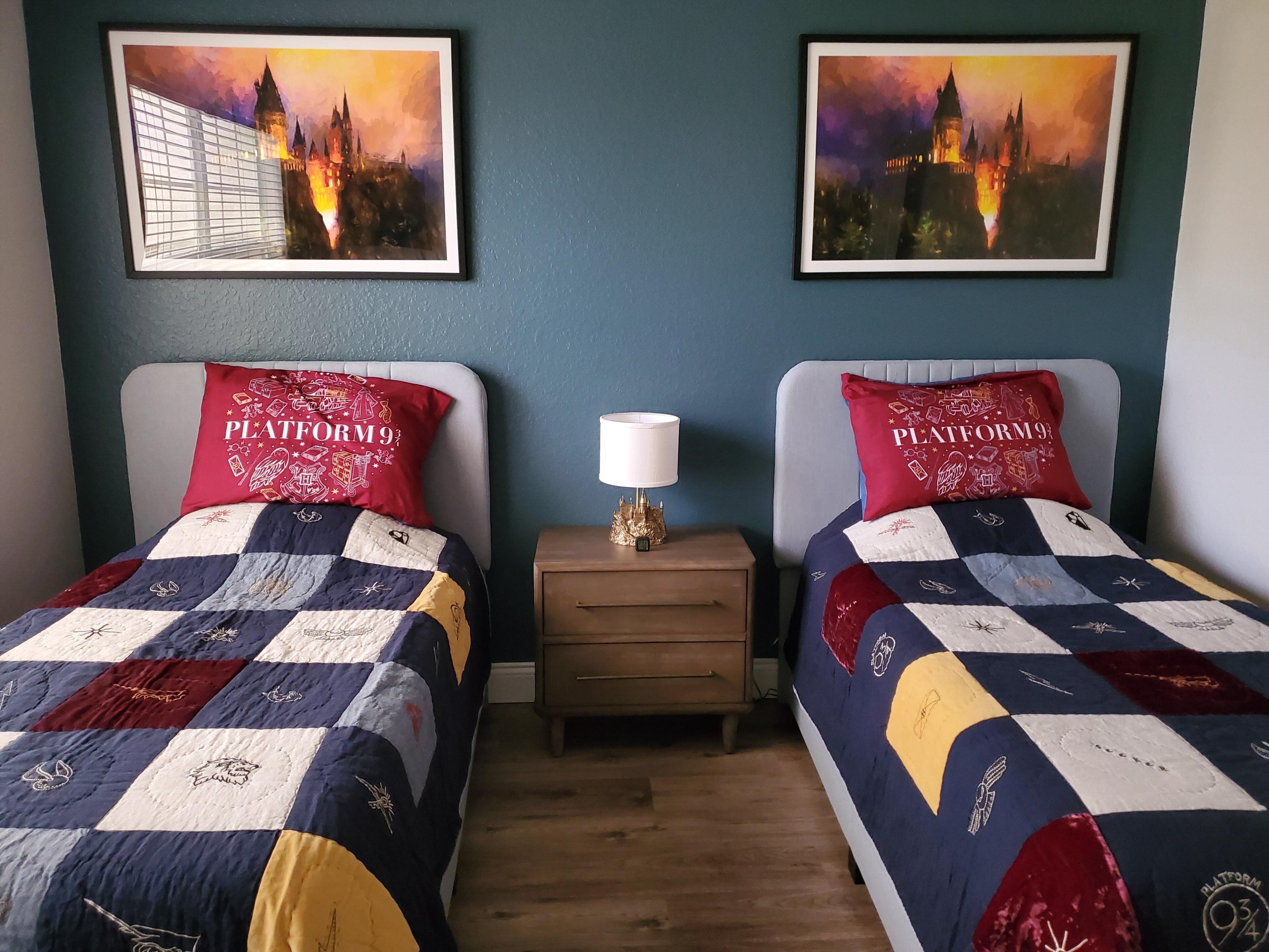 Une chambre avec deux lits, chacun orné d’un oreiller rouge sur lequel est inscrit « PLATE-FORME 9 ¾ ». Deux tableaux encadrés ornent le mur, ainsi qu’une table de chevet avec une lampe et une petite table en bois.
