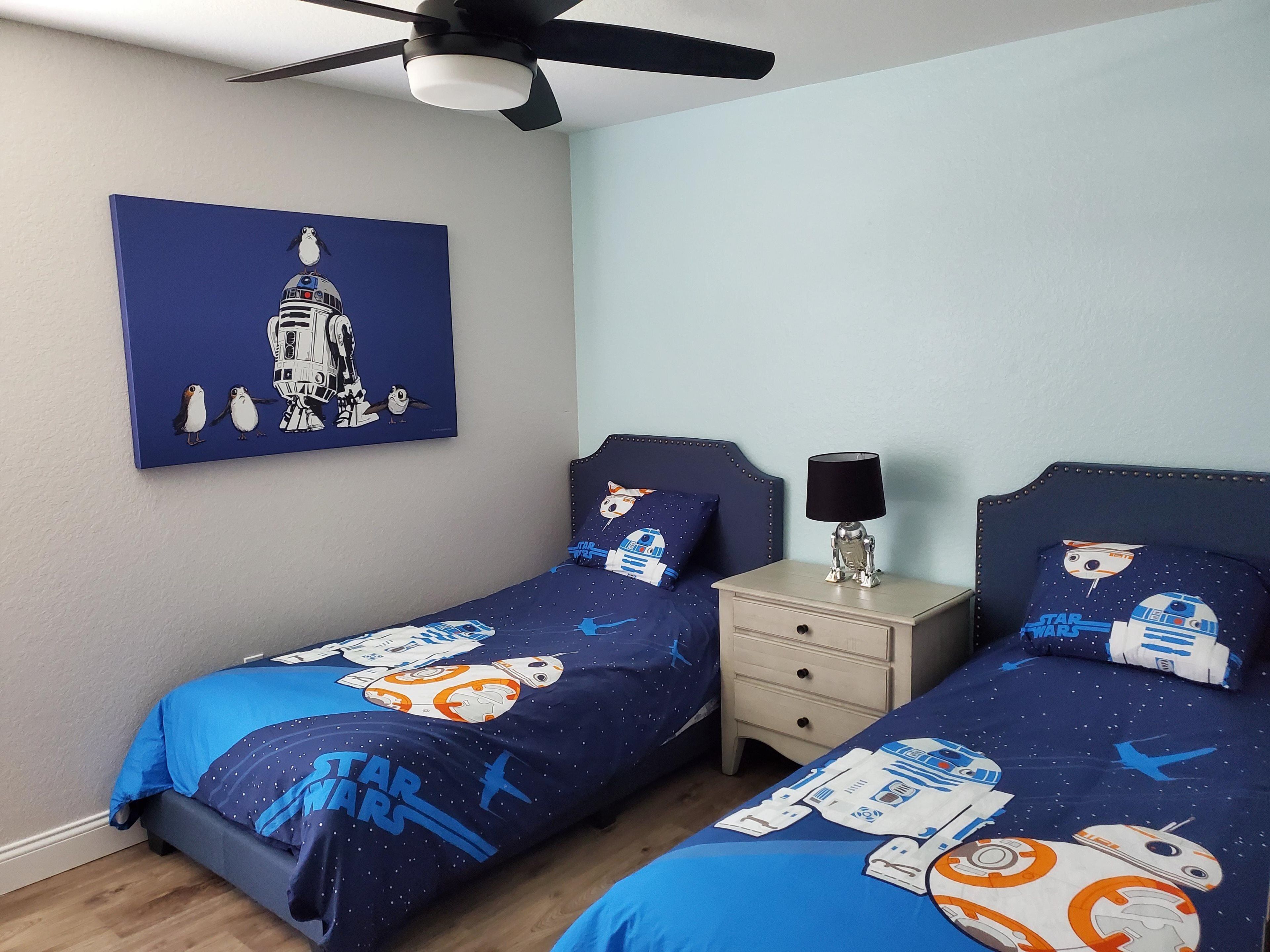 Une chambre avec deux lits, chacun agrémenté de draps et d’oreillers sur le thème de Star Wars, d’un ventilateur de plafond et d’une table de chevet avec une lampe.