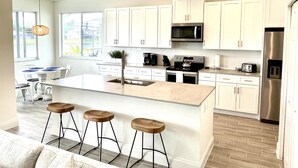 A modern kitchen with white cabinets, a central island with stools, stainless steel appliances, and a large window offering a view. - Lake House | Disney | Huge Pool&Spa | Gated (Kissimmee)