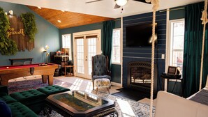 A cozy living room with a pool table, green sofas, a fireplace, and a television mounted on the wall. - 3+ acre Home: Pool Table, Putt-Putt, 4 miles to Downtown and SEMO, Summer Pool (Cape Girardeau)