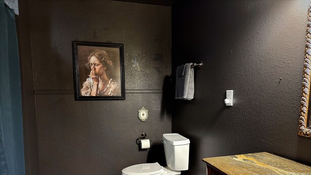 Une salle de bain avec un sol à carreaux noirs et blancs, des toilettes blanches, un lavabo avec un plan de travail en marbre et un tableau encadré au mur.